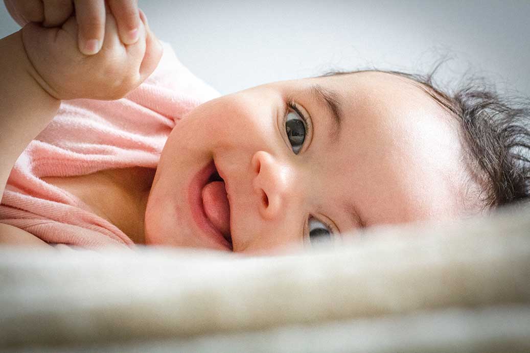 Photo of a smiling baby holding its hands lying in a bed.